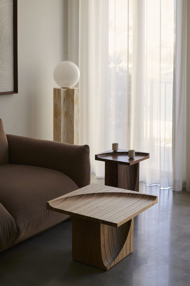 Minimalist living room with a brown sofa, two wooden tables, and a lamp on a wooden stand in front of sheer curtains