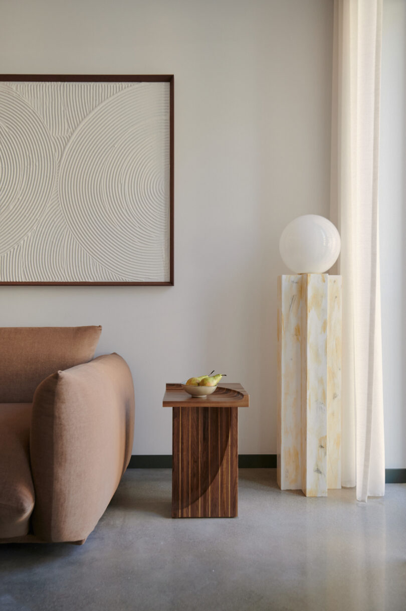 Modern living room with a brown couch, wooden side table with a bowl of fruit, abstract art on the wall, and a light-colored pedestal with a spherical lamp by the window