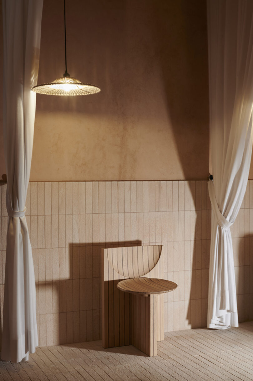 Minimalist interior with a wooden chair next to white curtains and a warm pendant light casting shadows on a textured beige wall