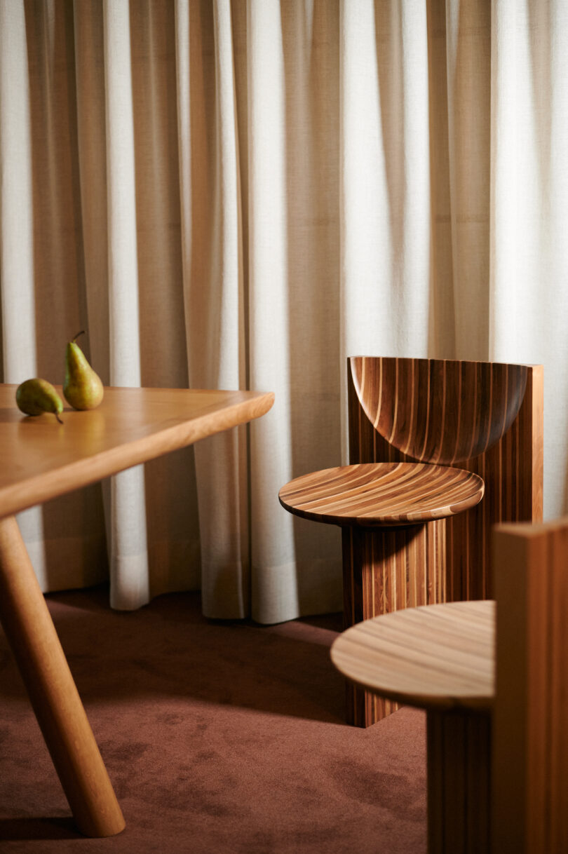 Minimalist wooden chairs and a table with pears on top, set against beige curtains on a brown carpet