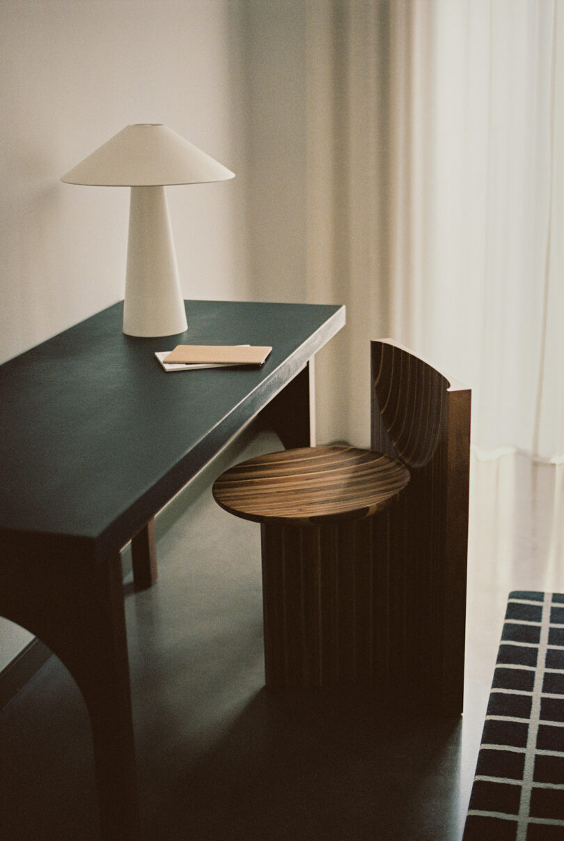 A minimalist workspace with a black desk, a white table lamp, and a wooden chair. A closed notebook and pen rest on the desk. Natural light filters through sheer curtains