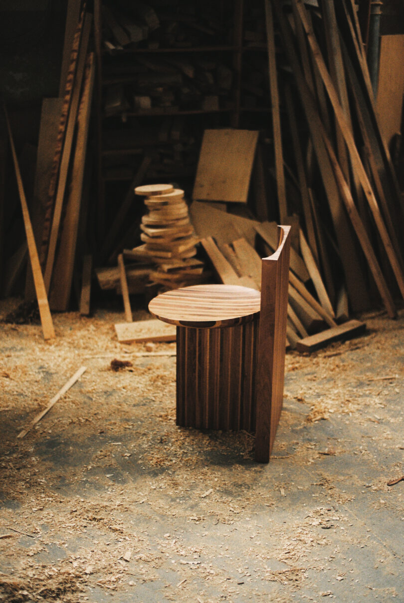 A wooden chair with a curved backrest stands in a workshop surrounded by scattered wood pieces and sawdust