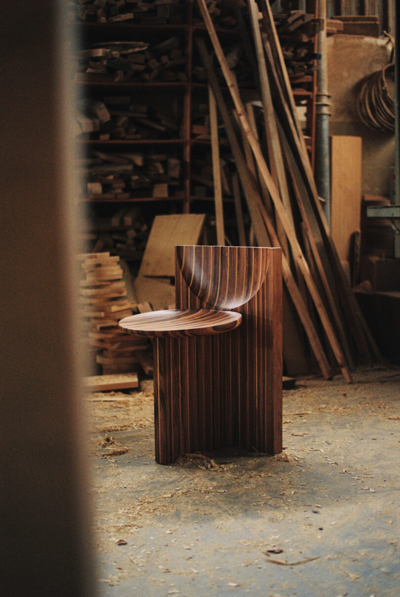 A wooden chair with a curved design is placed in a workshop. Various wooden planks and tools are visible in the background