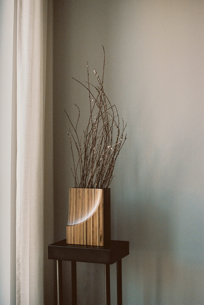 A wooden vase with branches on a small dark stand against a gray wall, next to a white curtain.