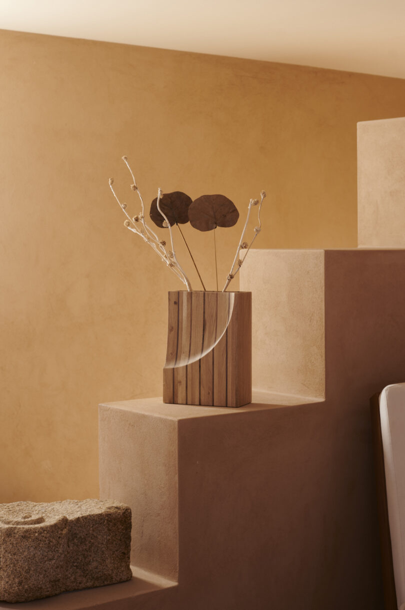 A wooden vase with dried botanicals is placed on a tiered, beige stone step against a light brown wall