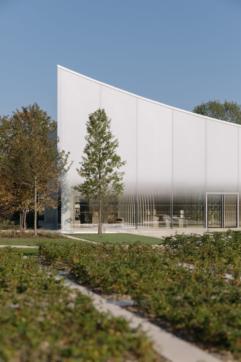 Sleek, modern building with a white facade featuring curved lines, large windows, and green landscaping in the foreground. Trees flank the structure under a clear blue sky.