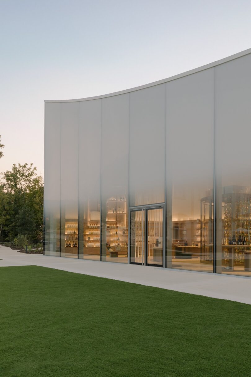 A modern building with a curved, translucent facade and visible interior lighting. The structure is surrounded by greenery and a concrete path.