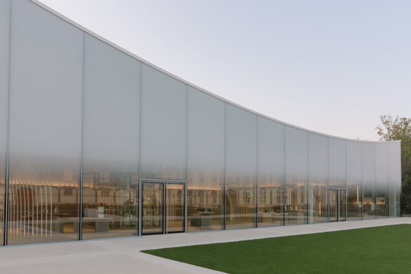 A modern building with a curved, translucent facade and a glass entrance, surrounded by grass under a clear sky.