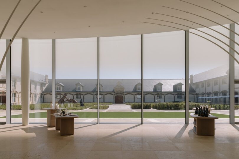 A spacious hall with floor-to-ceiling windows overlooks a modern courtyard with a landscaped garden. Inside, there are two tables displaying various decorative items.