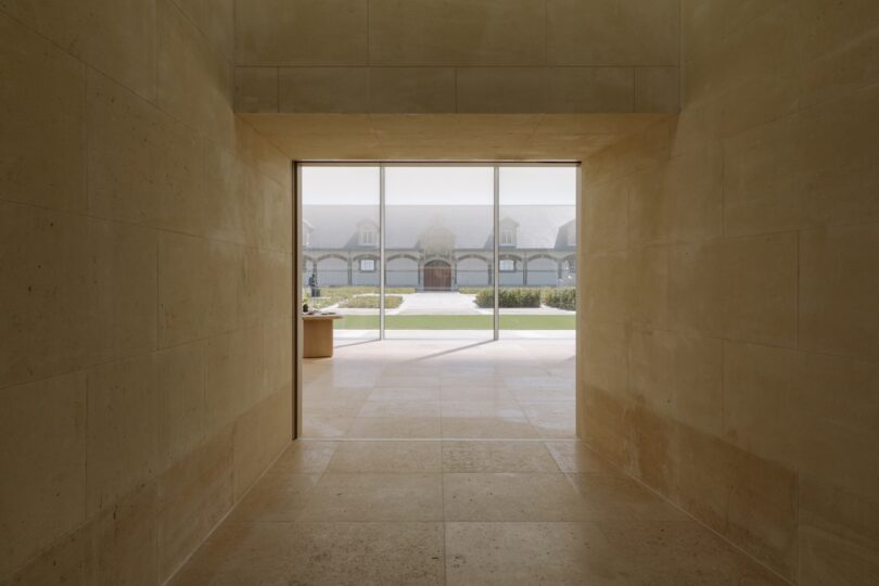 View through a stone hallway with a large square window, showing a courtyard and building with arched windows in the background.