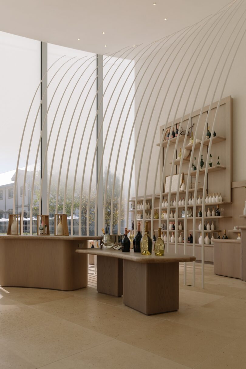 A modern store interior with wooden tables displaying bottles and items. Curved, white bars form an archway. Shelves of products line the wall. Large windows provide natural light.