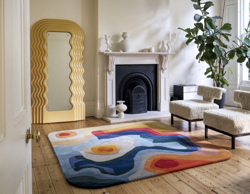 A stylish living room with a colorful abstract rug, two textured chairs, a wavy mirror, and a decorative mantle. A leafy plant stands near the window