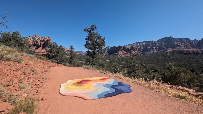 Abstract, wavy, colorful rug placed on a dirt path with a mountainous landscape in the background under a clear blue sky