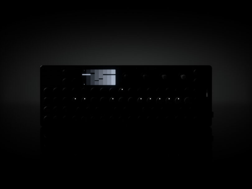 Dark image of a black electronic device with a small screen and multiple buttons, knobs, and indicators, partially illuminated against a black background.