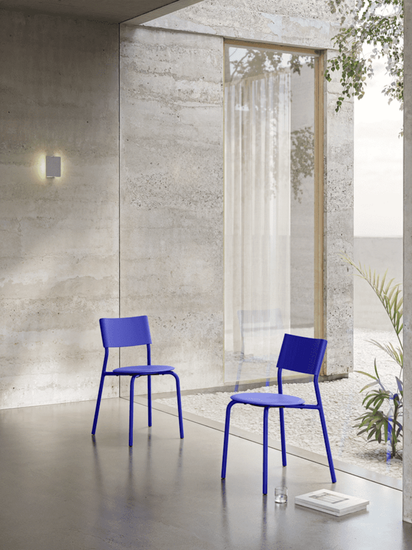 Two blue chairs in a modern room with concrete walls, a glass door, and a potted plant. A book and glass sit on the floor