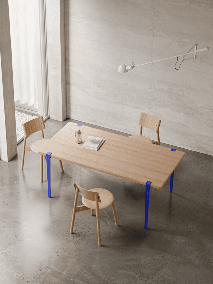 A minimalist dining room with a wooden table and three chairs on a concrete floor. Wall-mounted lamp above the table, a window with sheer curtains in the background