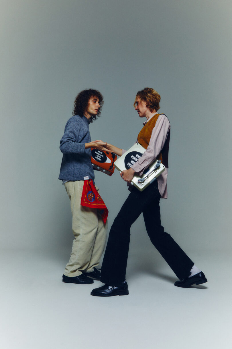 Two people stand facing each other, holding records and books. Both wear casual clothing and have long hair.