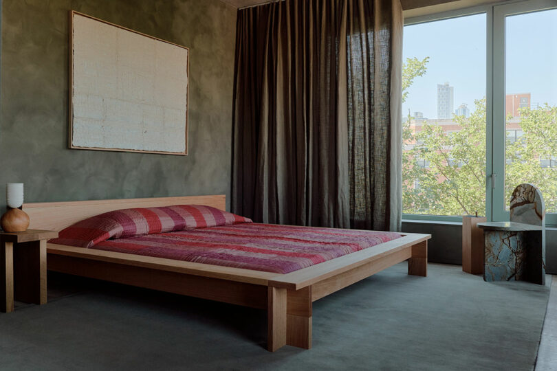 A modern bedroom with a low wooden bed, red striped bedding, green textured walls, large window with brown curtains, abstract art, and a small bedside table with a lamp.