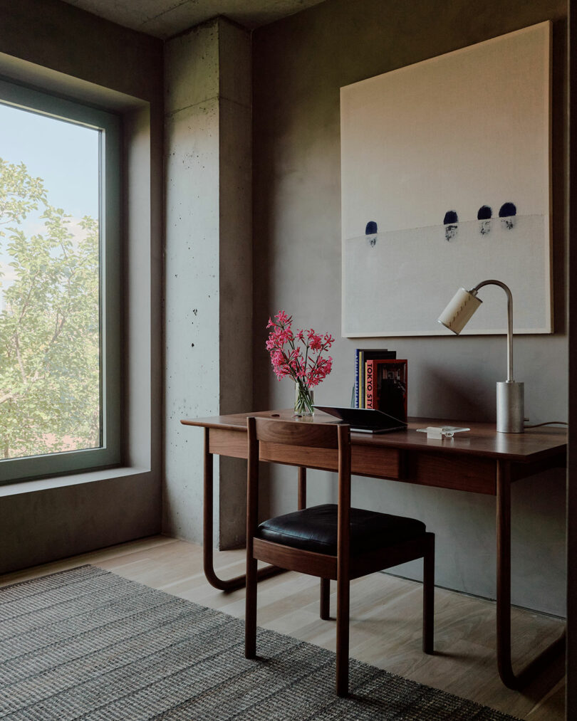 A minimalist office with a wooden desk, chair, lamp, red flowers in a vase, books, and a large abstract painting on the wall, near a window with a view of trees.