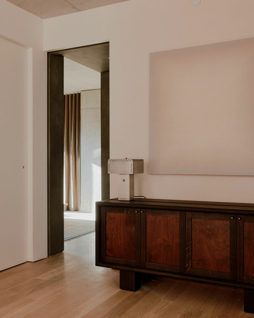 A minimalist room with a wooden sideboard, a modern lamp, and a large blank canvas on the wall. An open doorway leads to another room with curtains.