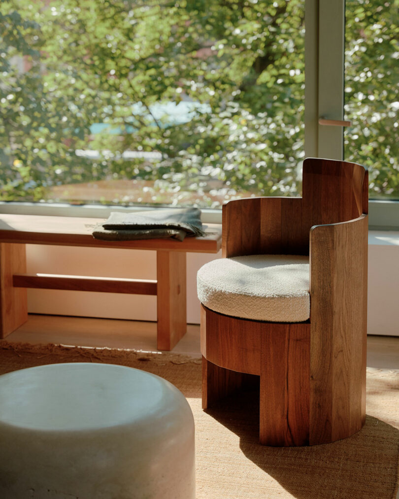 Wooden chair with cushioned seat by a window, next to a wooden bench with folded fabric and a round gray ottoman in front. Sunlit green leaves are visible outside.