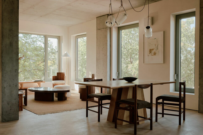 Modern dining and living area with large windows, wooden table, chairs, and a round coffee table. Neutral tones and natural light create a warm ambiance.