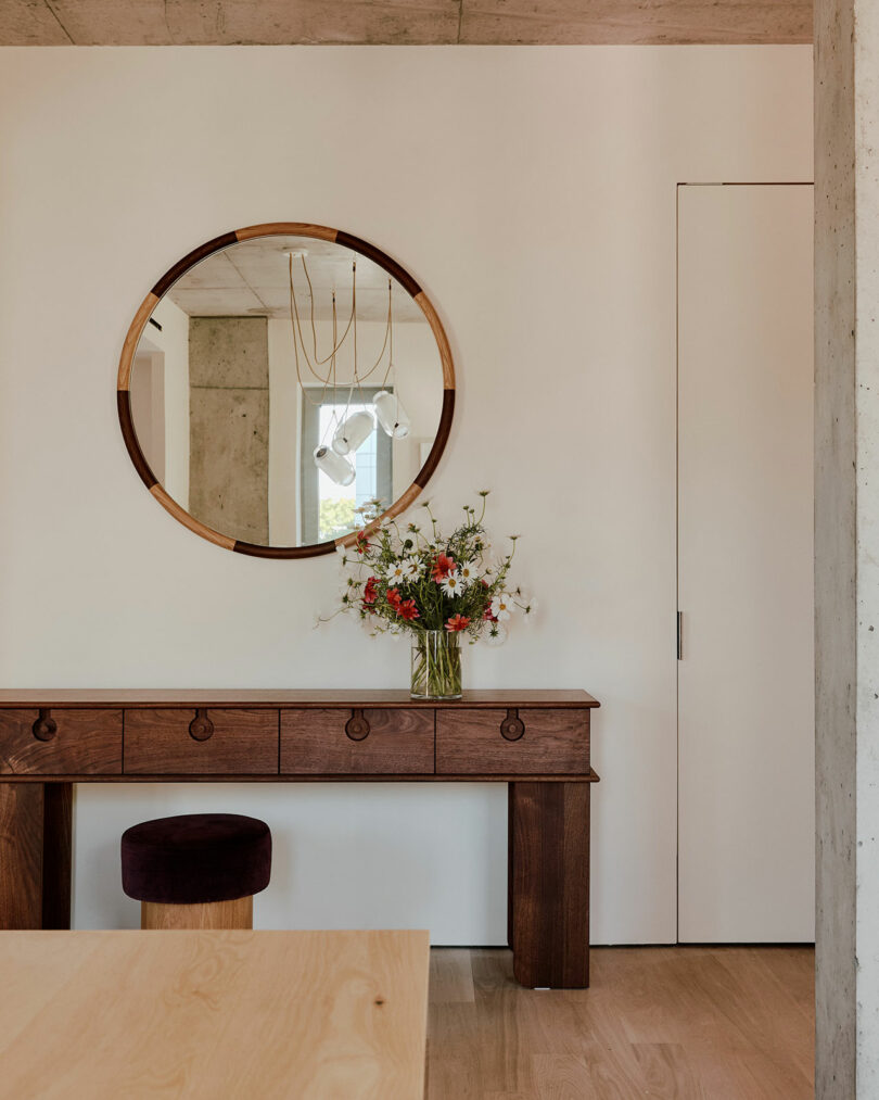 A wooden console table with a vase of flowers stands beneath a round mirror on a white wall. A small stool is tucked under the table.