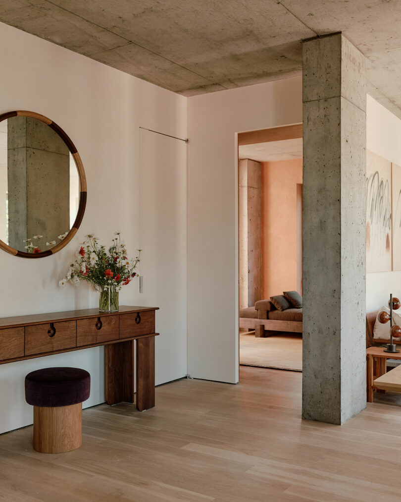 Interior with a wooden console table, round mirror, and stool. A concrete pillar and doorway lead to a living area with a sofa. Flowers are placed on the table.