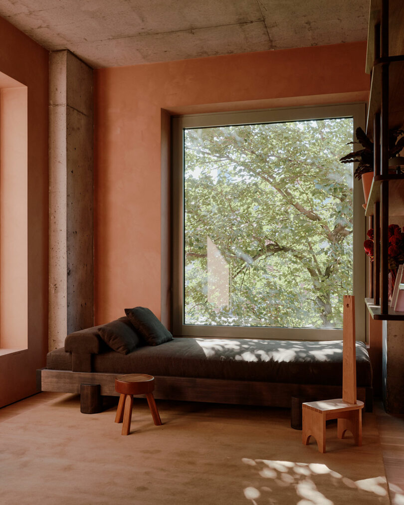 A cozy room with a brown daybed near a large window, offering a view of green trees. Two wooden stools are positioned by the daybed, and shelves hold decorative items on the right.