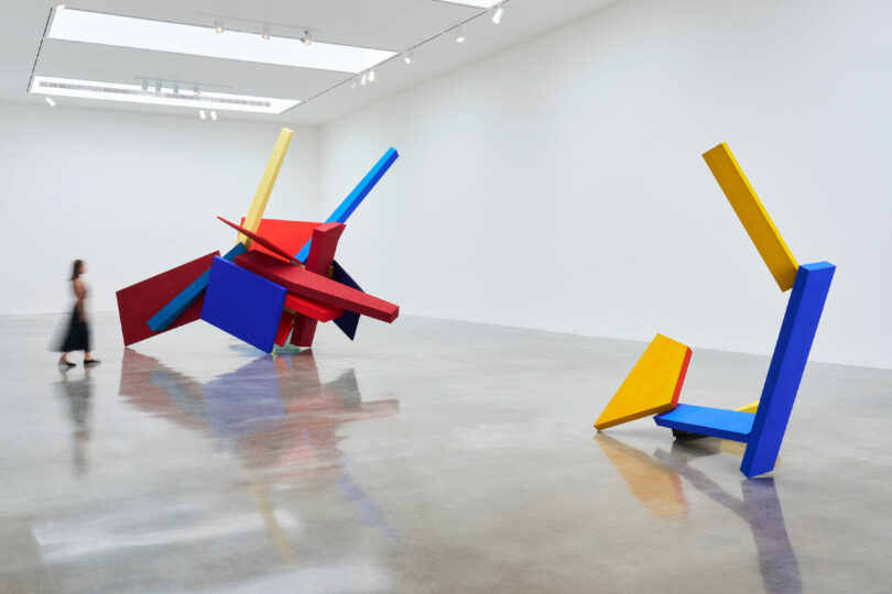 A person walks near large geometric sculptures in red, blue, and yellow in a modern, white-walled gallery space with polished concrete floors.