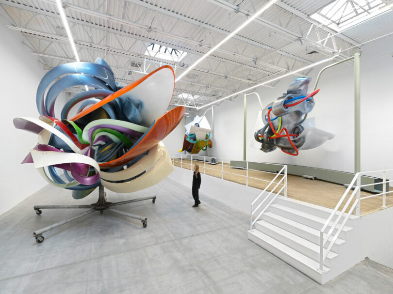 A person stands in a gallery with large, colorful abstract sculptures under a high ceiling with skylights.