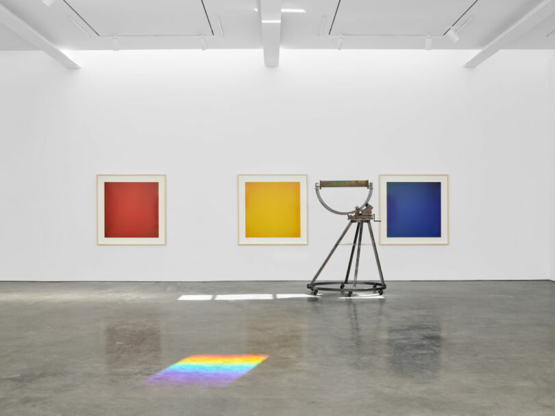 Art gallery with three framed, solid-colored gradients on the wall: red, yellow, and blue. A metallic sculpture stands on a polished concrete floor with a rainbow light reflection.