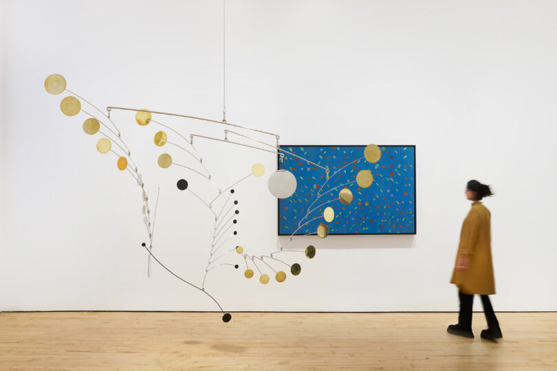 A person in a brown coat walks past a large hanging mobile sculpture and a framed blue artwork on a gallery wall.