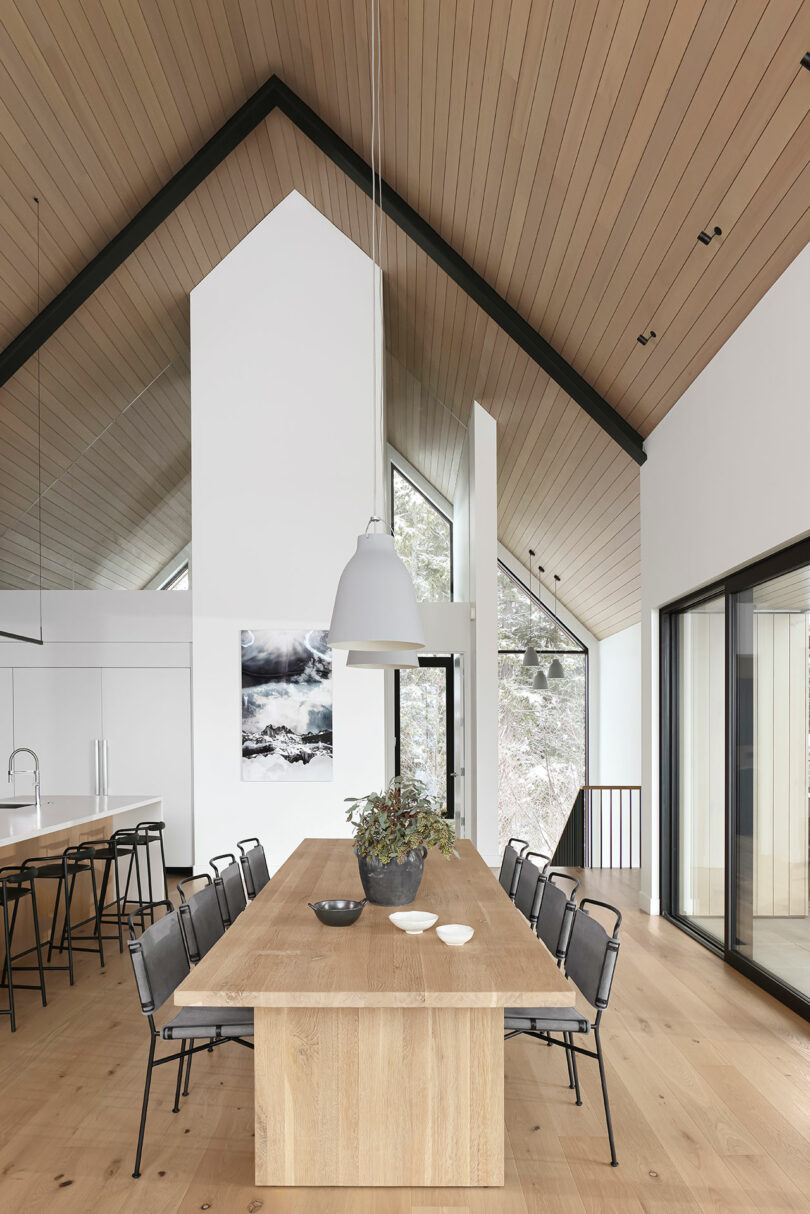 Comedor moderno con una larga mesa de madera, sillas negras, iluminación colgante y grandes ventanales con vistas a un paisaje cubierto de nieve.