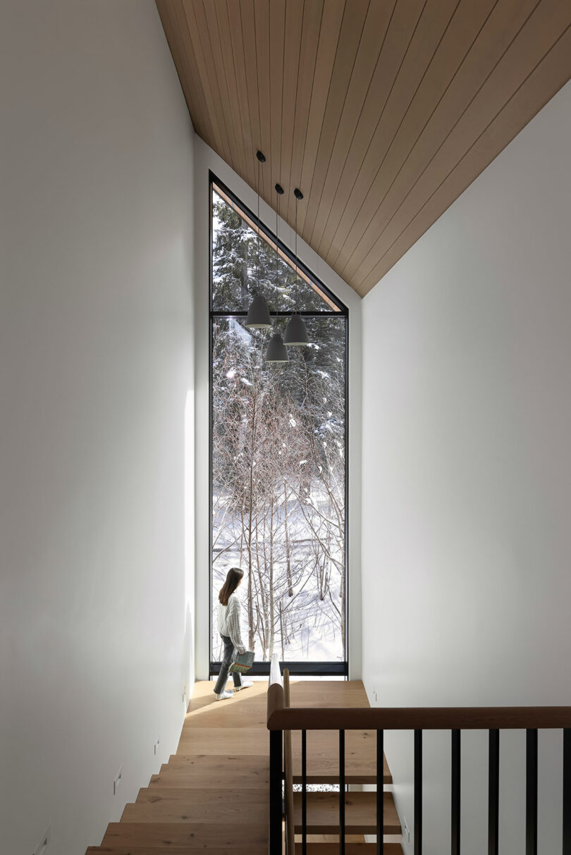 Una persona caminando por una escalera de madera junto a una gran ventana con una vista exterior nevada.