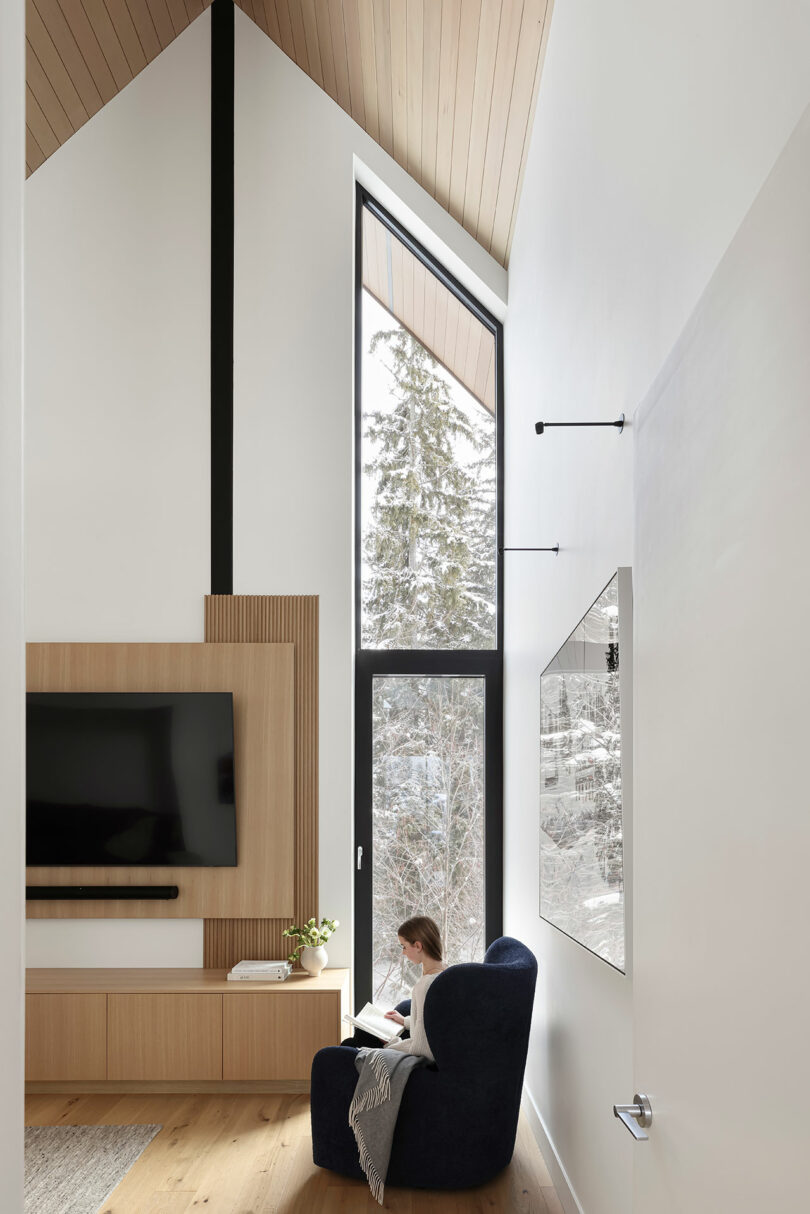 Una persona sentada en una silla negra lee junto a un gran ventanal en una sala de estar moderna. La habitación cuenta con un panel de TV de madera y techos altos con detalles en madera. Afuera se ven árboles nevados.