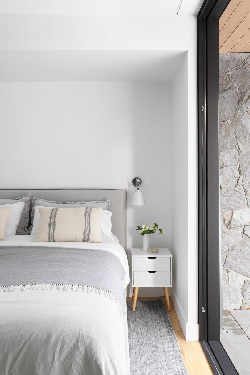 Dormitorio minimalista con cama, ropa de cama blanca y gris, mesita de noche con lámpara y planta, junto a una gran puerta corredera de cristal que da a una pared de piedra.