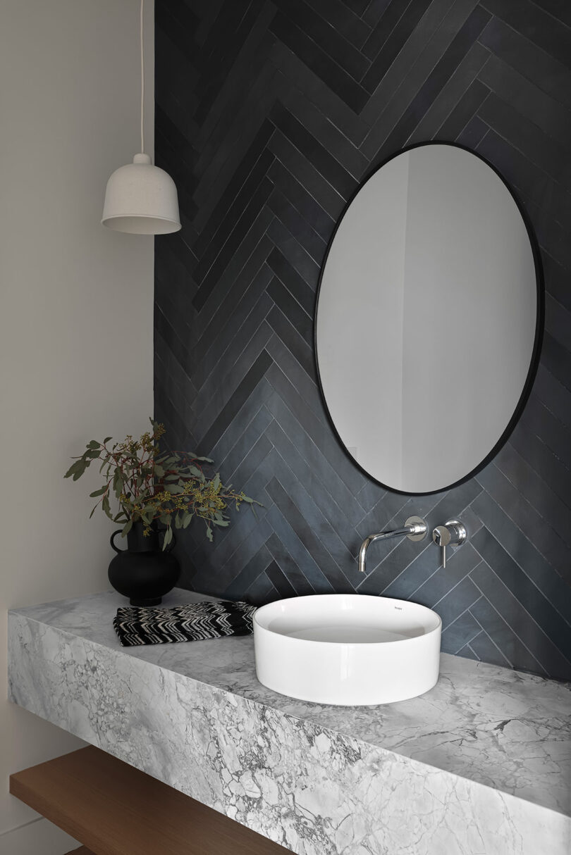 Cuarto de baño con espejo redondo, pared de azulejos en forma de espiga, lámpara colgante blanca, encimera de mármol, lavabo blanco redondo y jarrón negro con vegetación.