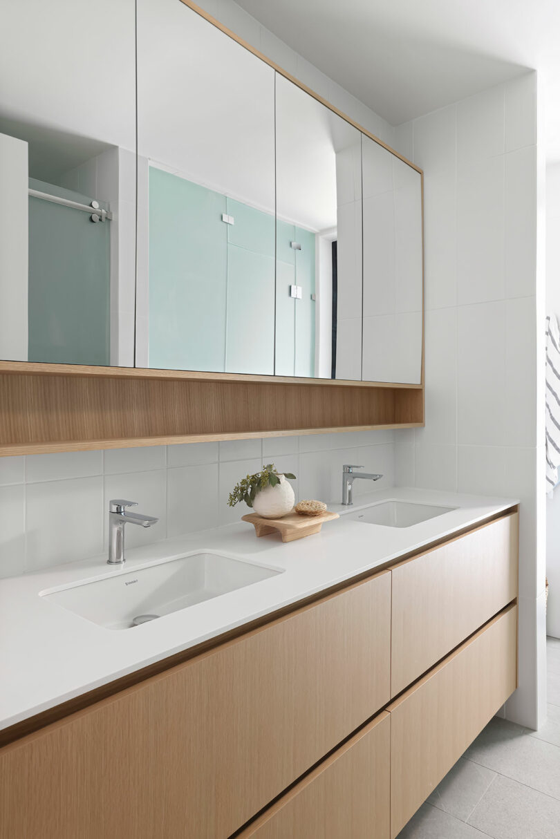 Baño moderno con tocador de doble lavabo, gabinetes de madera, espejos grandes y una planta decorativa. El suelo es de color gris claro y las paredes están alicatadas en blanco.