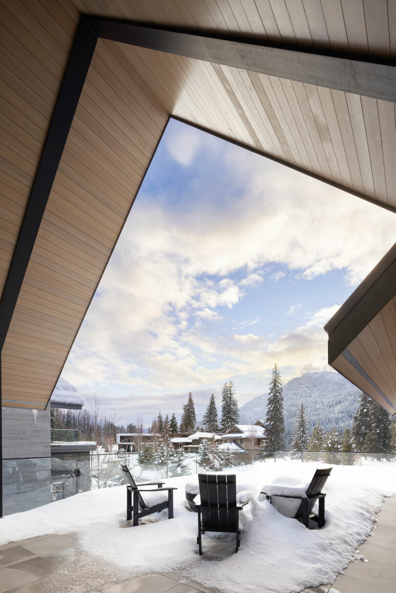 Patio nevado con tres sillas vacías bajo un moderno techo de madera. Al fondo se ven árboles y montañas cubiertos de nieve bajo un cielo parcialmente nublado.