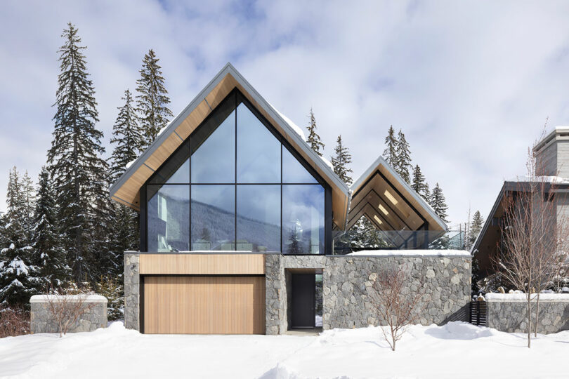 Casa moderna con grandes ventanales de cristal, paredes de piedra y detalles en madera, rodeada de nieve y árboles de hoja perenne.