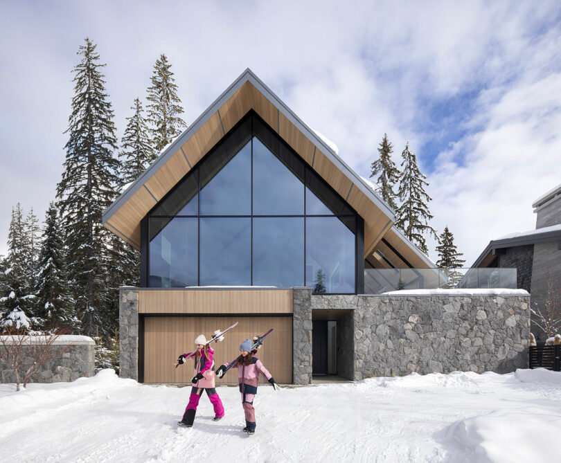 Dos personas vestidas de invierno llevan esquís frente a una casa moderna en forma de A con grandes ventanales de vidrio, rodeada de nieve y árboles altos.