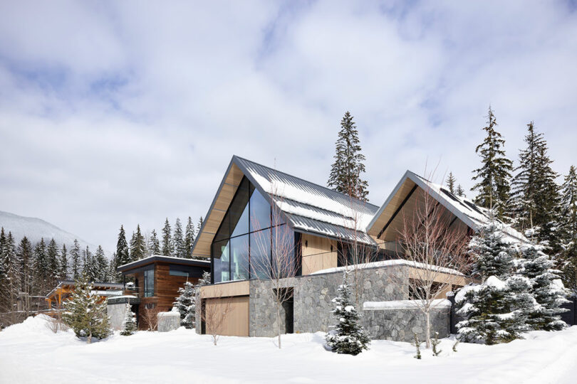 Casa moderna con grandes ventanales y fachada de piedra rodeada de nieve y pinos bajo un cielo nublado.