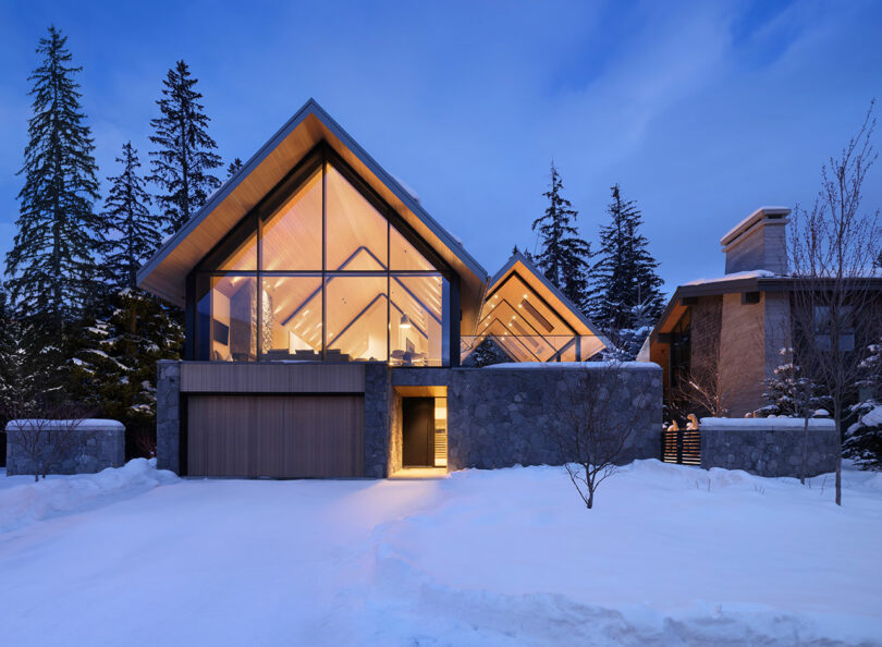 Una casa moderna con grandes ventanales de cristal y techo inclinado está rodeada de nieve y árboles altos bajo un cielo crepuscular. La iluminación interior aporta a la casa un brillo cálido.