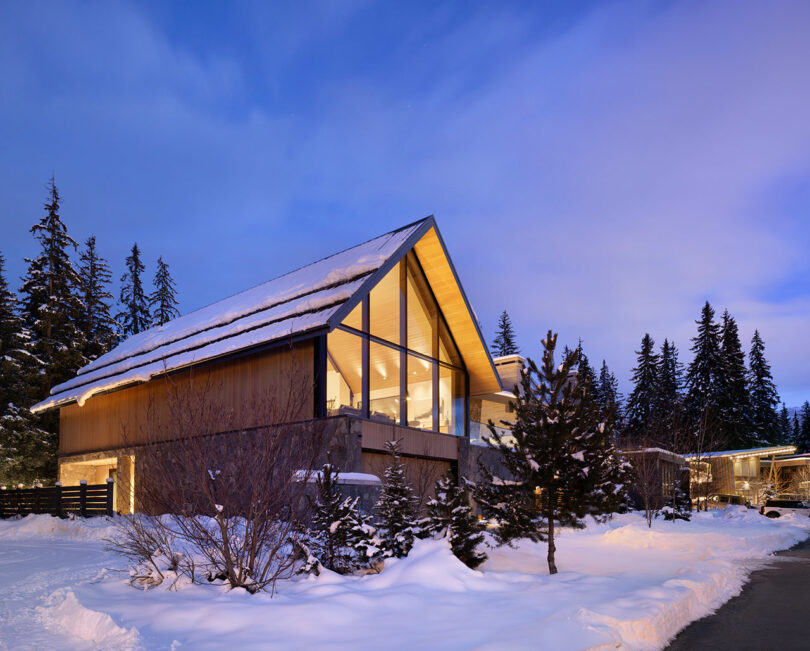 Una casa moderna cubierta de nieve con grandes ventanales de cristal y un techo empinado, rodeada de pinos durante el crepúsculo.