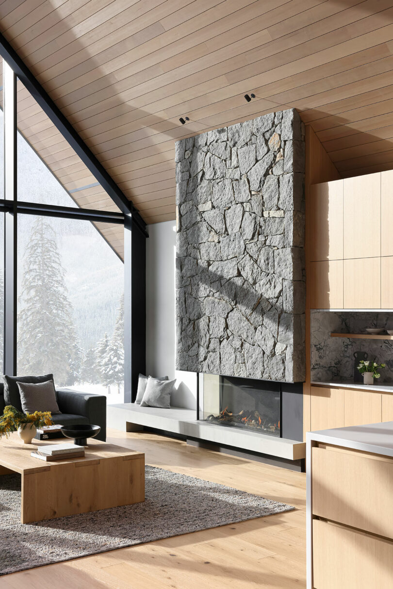 Salón moderno con una gran chimenea de piedra, techo de madera y grandes ventanales que muestran un paisaje nevado. Muebles y decoración en tonos neutros completan el espacio.