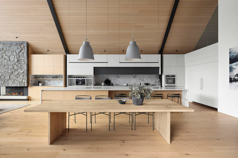 Cocina moderna con una gran mesa de comedor de madera, sillas negras, luces colgantes y chimenea de piedra. Los gabinetes blancos y el protector contra salpicaduras de mármol realzan el diseño elegante. .