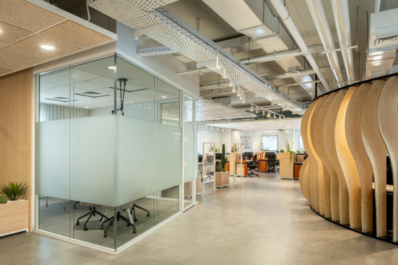 Espace de bureau moderne avec salle de réunion aux parois de verre, espace de travail ouvert et cloisons incurvées en bois. Le plafond comprend des tuyaux et un éclairage apparents. Les plantes décorent l'espace.