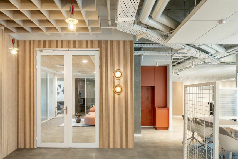 Espace de bureau moderne avec plafond ouvert, tuyaux apparents, panneaux de bois, portes vitrées et mobilier minimaliste.