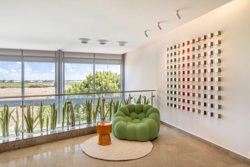 A modern seating area with a green armchair, small orange table, and checkerboard art on the wall. Large windows provide a view of the outdoors, with plants lining the glass railing.